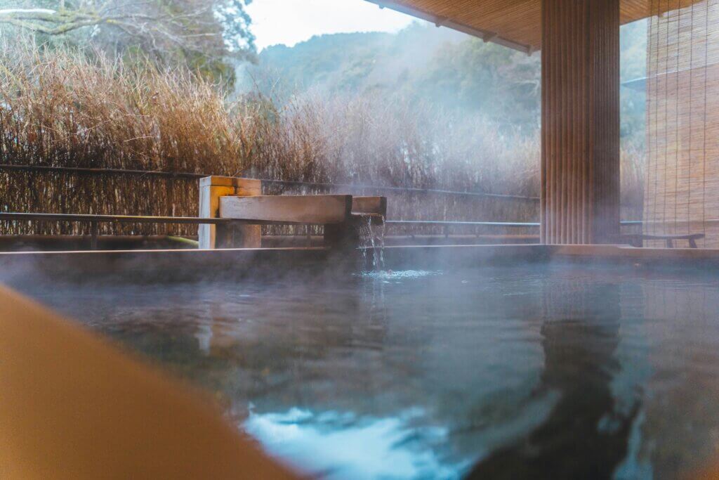 A traditional Japanese onsen bath with wooden tubs, steaming mineral water, and a tranquil mountain backdrop.