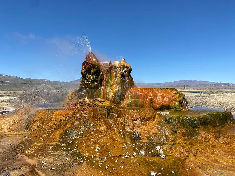 Small geothermal geyser in Nevada spraying water, covered in bright red, green, and yellow mineral deposits.